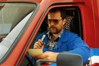 Ein Mann in blauer Arbeitskleidung sitzt am Steuer eines roten Fahrzeugs und liest konzentriert einen Zettel, den er hält.