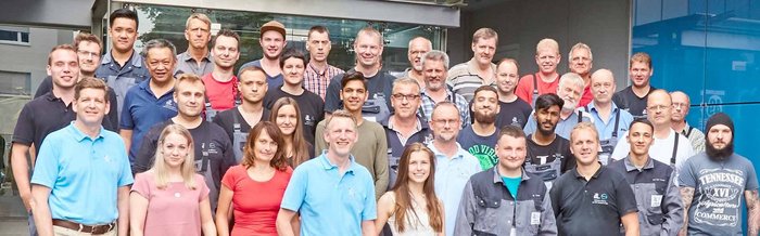 Die Gruppe besteht aus Männern und Frauen unterschiedlichen Alters, die vor einem modernen Gebäude stehen und in die Kamera lächeln.