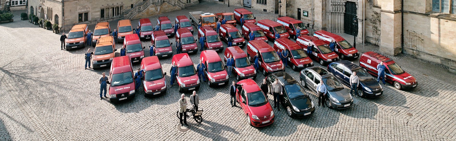 Zahlreiche rote Lieferwagen und einige graue Autos sind auf einem historischen Platz versammelt, umgeben von Mitarbeitern.