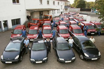 Eine Gruppe von Mitarbeitern steht stolz vor einer Flotte roter und schwarzer Lieferwagen auf einem gepflasterten Platz.