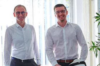 Zwei Männer in weißen Hemden stehen neben einem alten Fahrrad mit dem Namen 'W. Ahlers' auf einem hellen Boden.