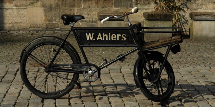 Das Fahrrad mit der Aufschrift 'W. Ahlers' hat ein einzigartiges Design und steht vor einer steinernen Wand.