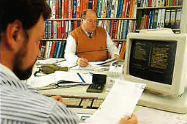 Im Büro sind zwei Männer zu sehen, einer liest Unterlagen, der andere konzentriert sich auf den Bildschirm eines alten Computers.